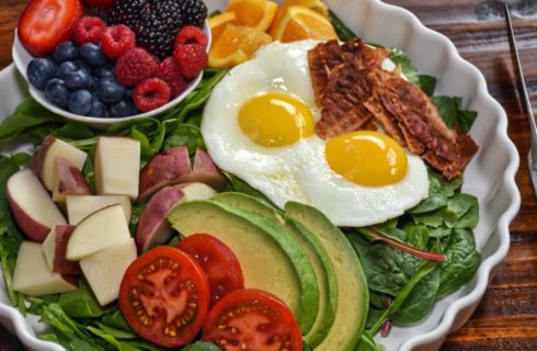 Breakfast dish full of fresh greens, sliced avocados and tomatoes, potatoes, overeasy eggs, bacon, strawberries, blueberries, raspberries, and blackberries