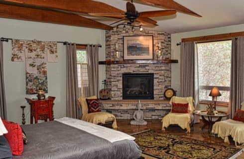 Large suite with bed and gray bedding, stone fireplace, multiple chairs, large area rug, and large windows