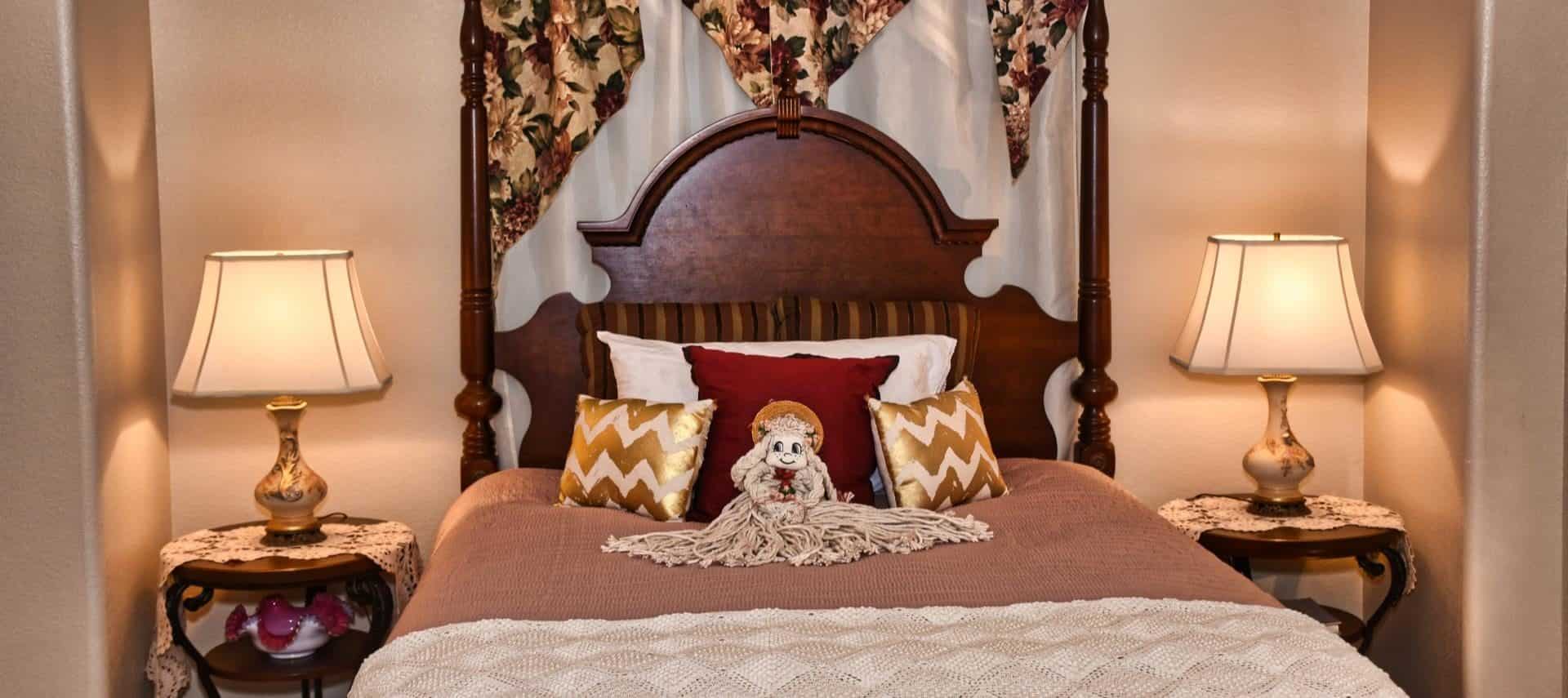 Bedroom with large dark wooden headboard with neutral bedding, white, red, and gold pillows, mop doll, and end tables with lamps